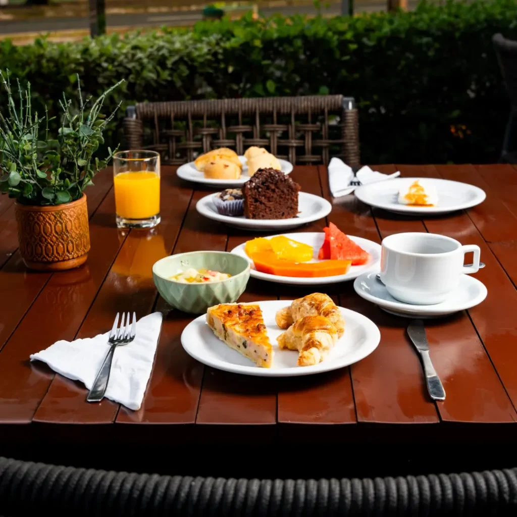 Mesa de madeira com café da manhã: quiche, croissant, frutas, salada de frutas e xícara de café.