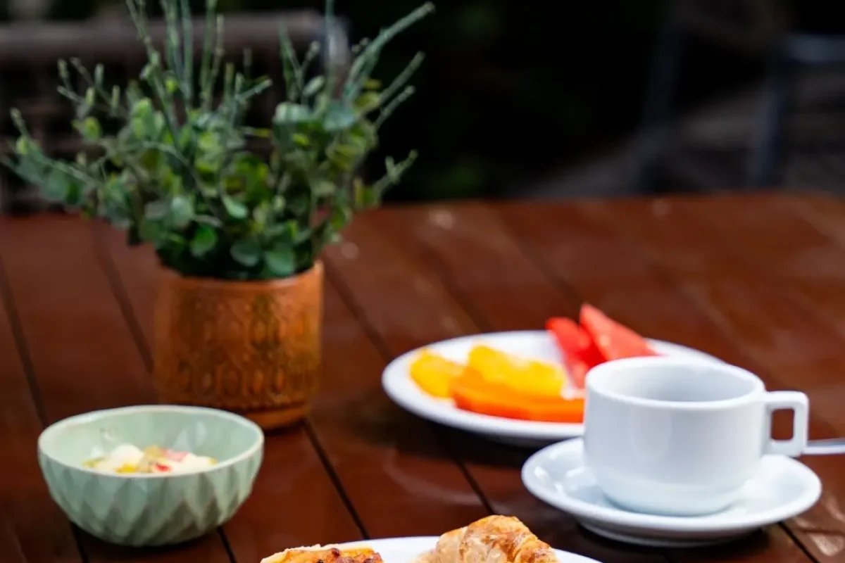 Mesa de madeira com café da manhã: quiche, croissant, frutas, salada de frutas e xícara de café.